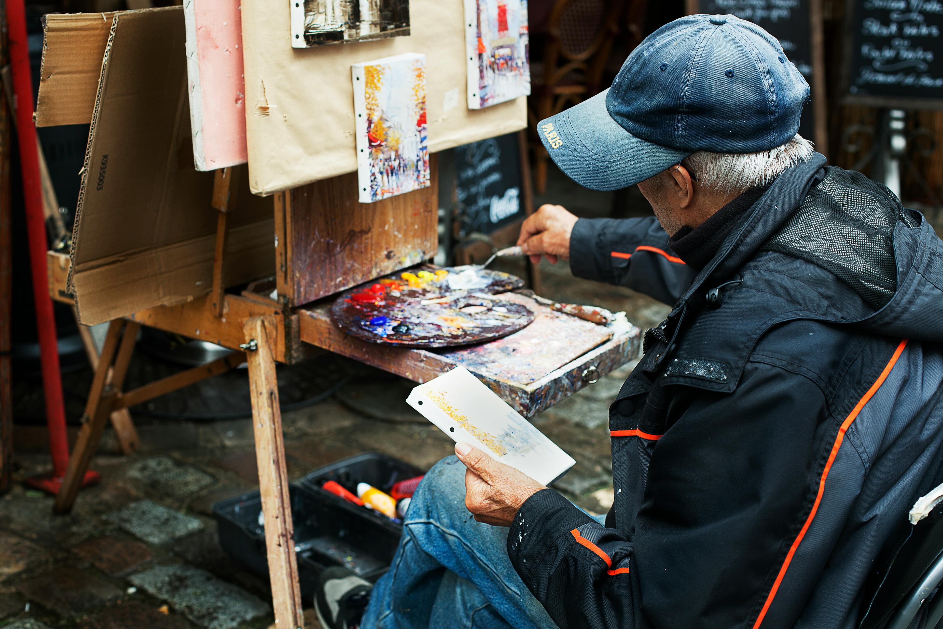 An elderly artist sits in front of an easel, painting on a small canvas while surrounded by vibrant paint tubes and a palette.