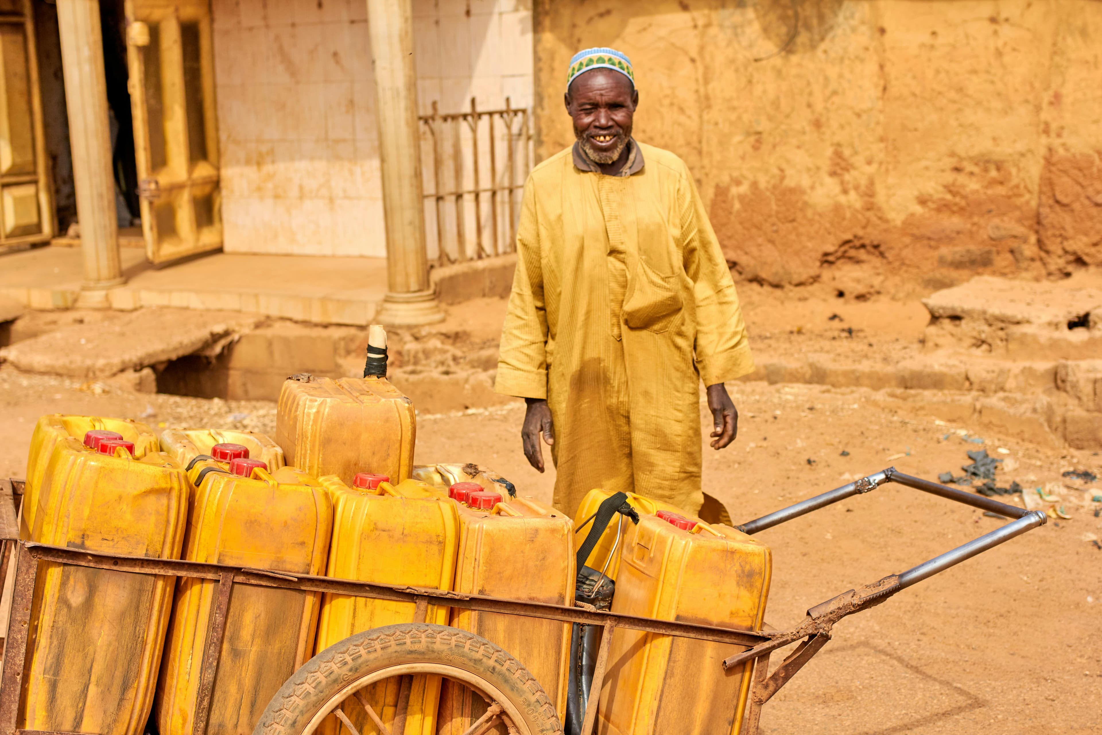 A person in a yellow robe stands beside a wooden cart filled with yellow jerry cans, set against a dusty street background.