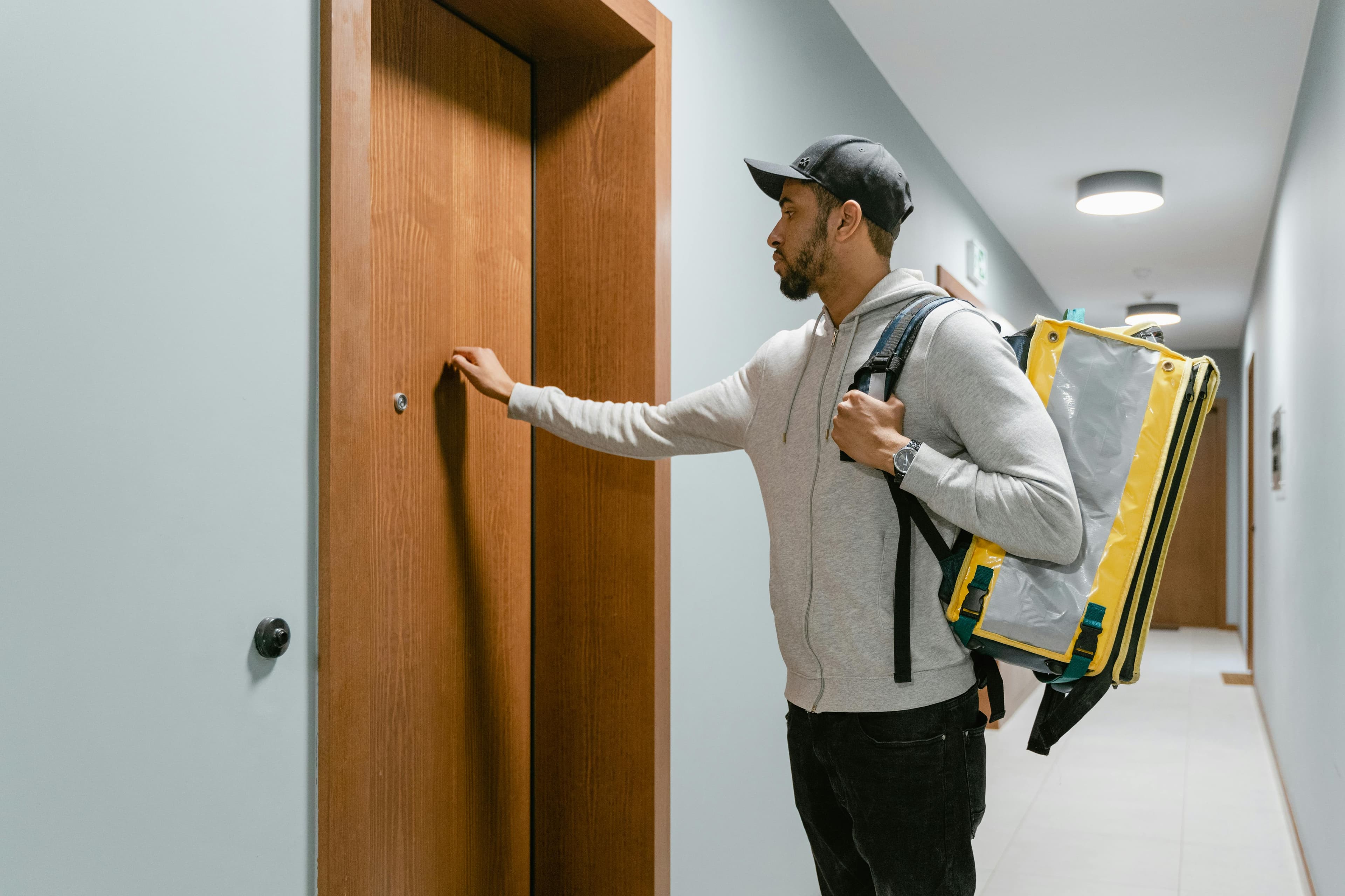A delivery person stands in a hallway, reaching to knock on a wooden door while carrying a yellow insulated bag on their back.