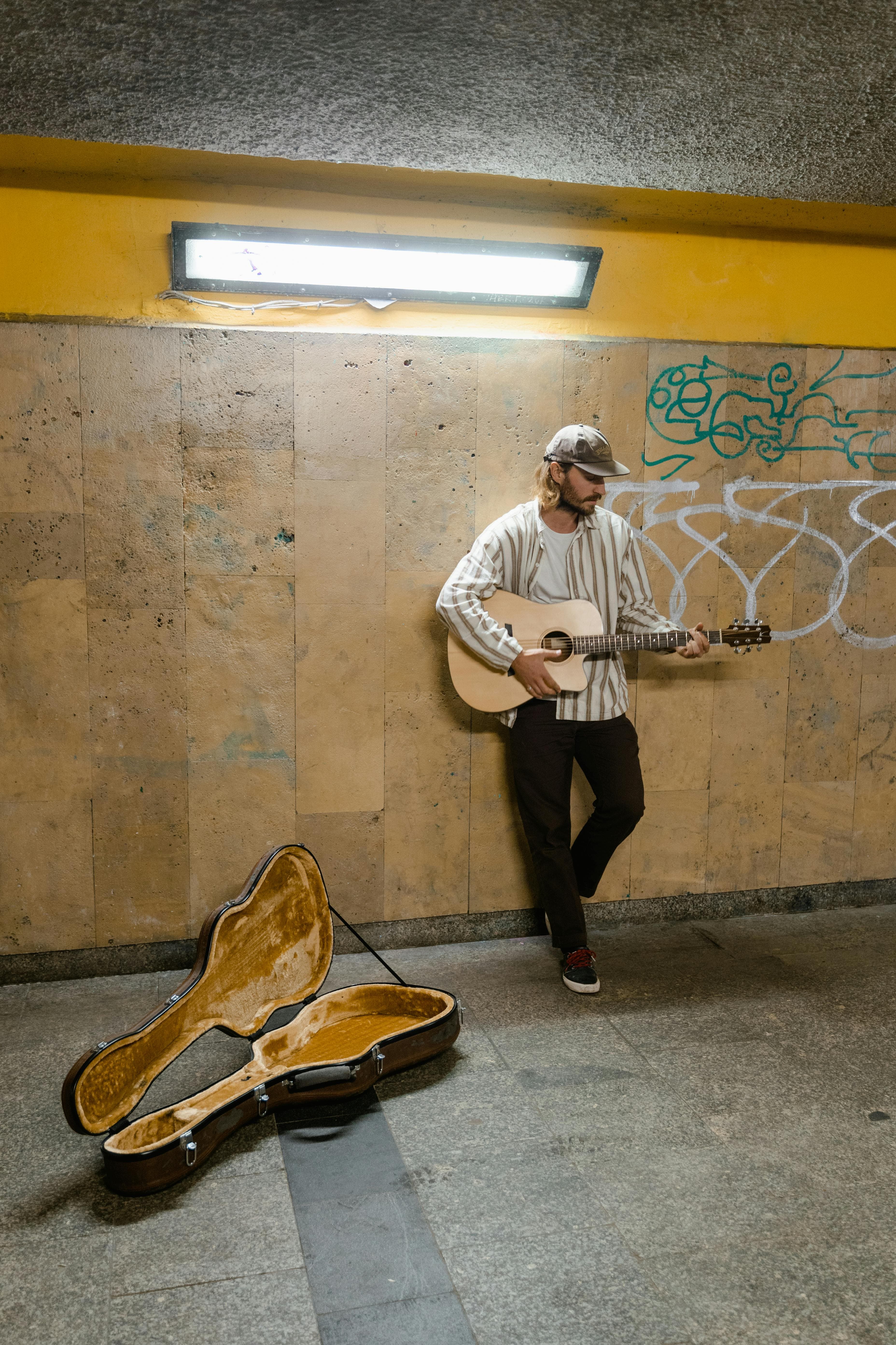A musician stands in a subway station, playing guitar beside an open case on the ground, with graffiti on the walls behind.