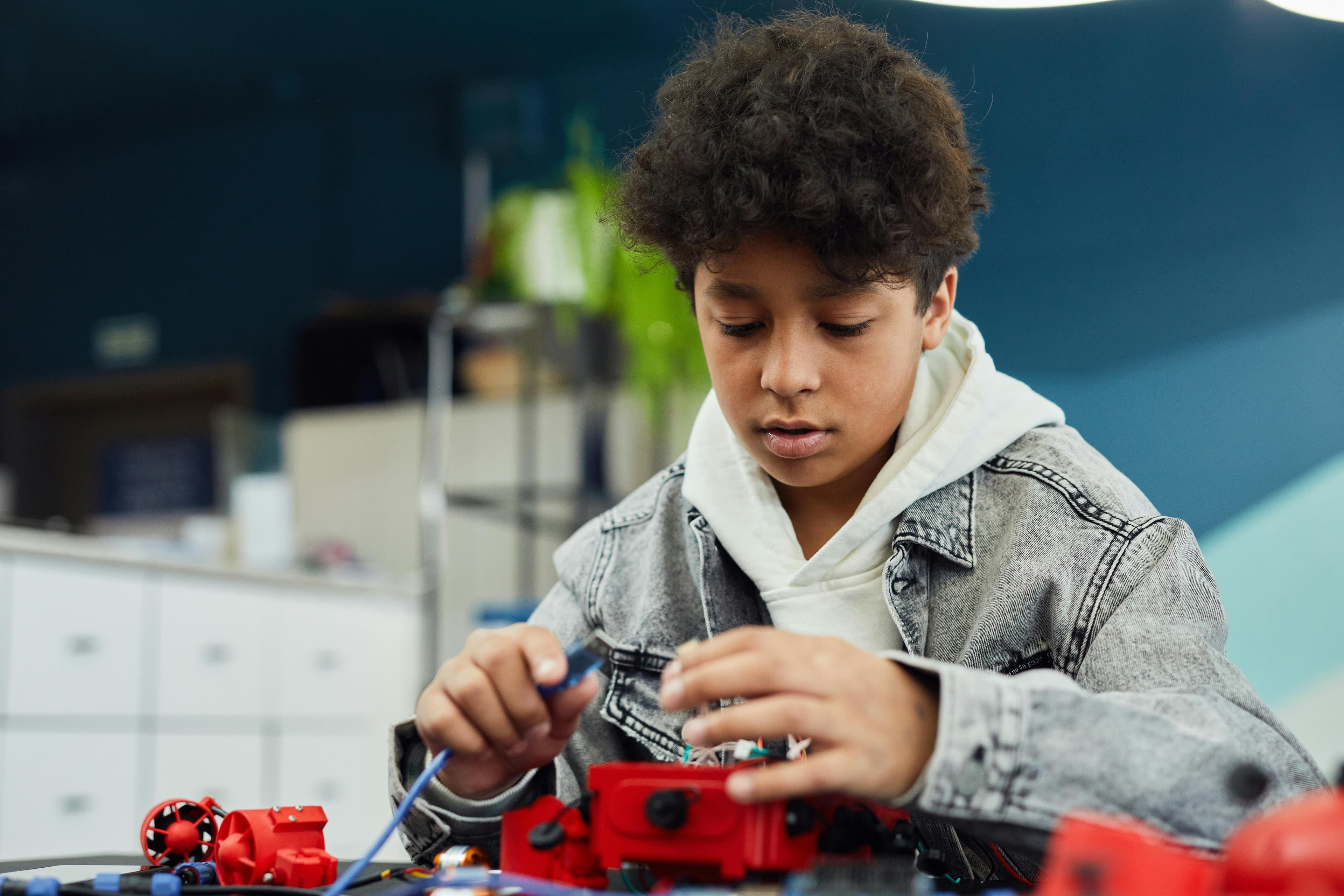 A person with curly hair wearing a denim jacket works intently on red robotic components, surrounded by tools and a colorful workspace.