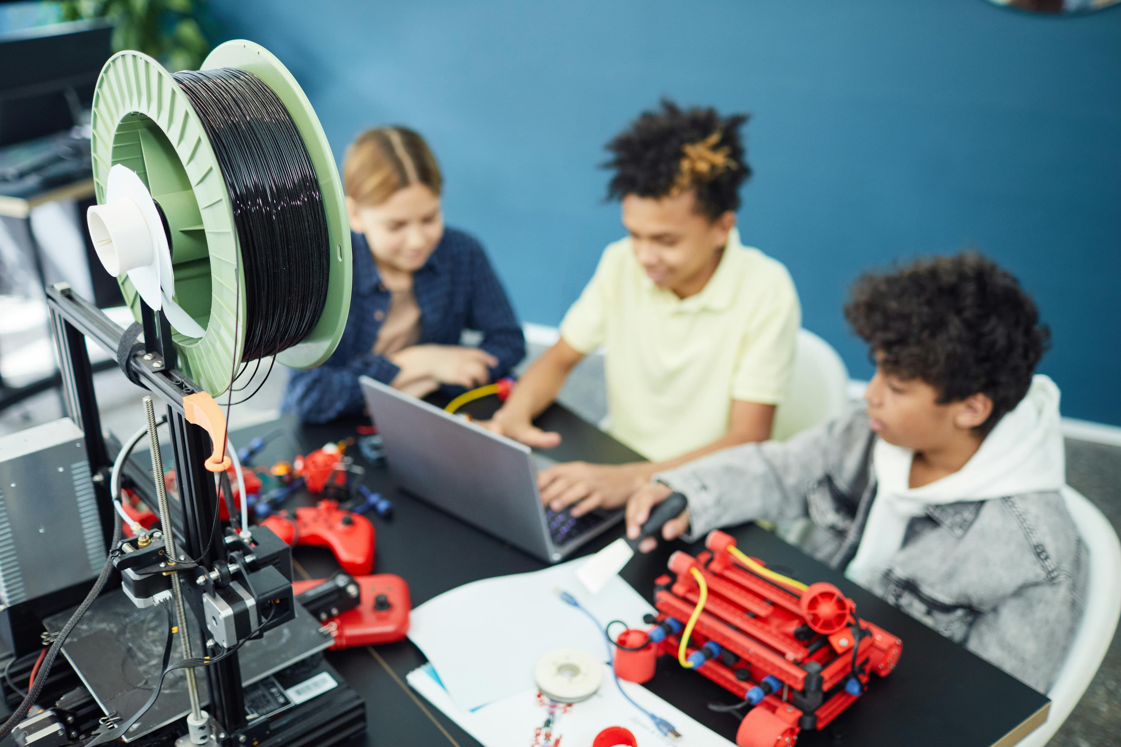 A 3D printer with filament stands beside children engaged in a tech project, working on laptops and using robotics components.